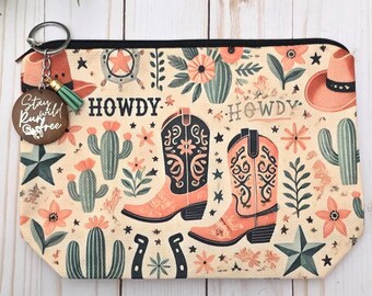 May include: A beige zippered pouch with a Western-themed design. The pouch features illustrations of cowboy boots, cacti, stars, and the word "HOWDY". A keychain with a wooden tag that says "Stay Wild Run Free" is attached.