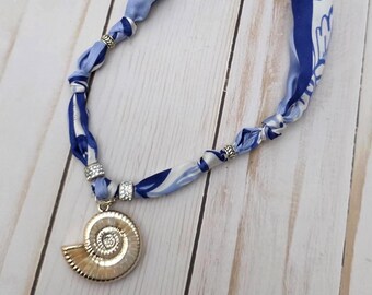 May include: A handmade necklace featuring a gold seashell pendant with a spiral design. The necklace's cord is made of blue and white patterned fabric, accented with silver-toned beads. The necklace is approximately 18 inches long.