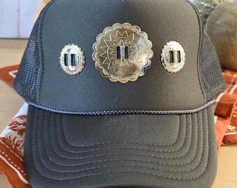 May include: A dark gray trucker hat with a mesh back and a curved brim. The hat features three silver-colored decorative conchos. The hat is sitting on a patterned orange and white bandana.