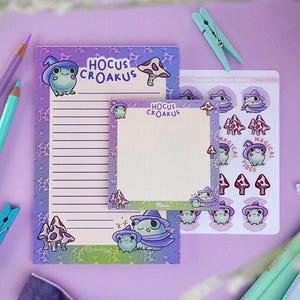 May include: A stationery set featuring a whimsical frog and mushroom design with the text "Hocus Croakus." The set includes a lined notepad, a blank notepad, and a sheet of stickers. The color scheme is purple, green, and white.