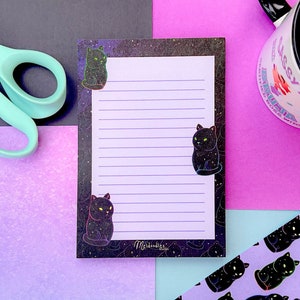 Puede incluir: Se muestra un bloc de notas con papel rayado e ilustraciones de gatos negros, un marcapáginas a juego y una taza con el texto "Messy Miss". También se ven unas tijeras verde menta. El bloc de notas y el marcapáginas presentan un diseño de noche estrellada.