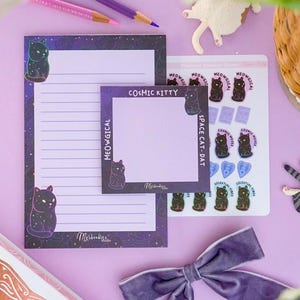 May include: A collection of stationery items with a cosmic cat theme, including notepads, stickers, and pencils. The notepads feature a purple galaxy design with a black cat illustration. The stickers have cat and celestial designs. A purple velvet bow adds a decorative touch.