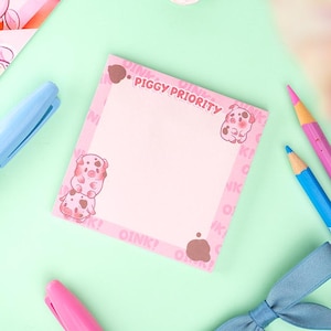 Puede incluir: Un bloc de notas rosa con el texto "PIGGY PRIORITY" e ilustraciones de cerdos. El bloc de notas est&aacute; rodeado de l&aacute;pices rosas y azules, un rotulador rosa y un lazo azul sobre una superficie verde claro. Papeler&iacute;a.