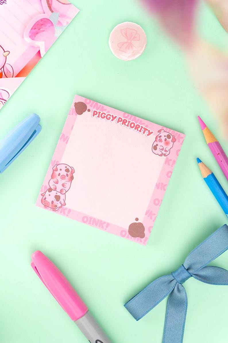 Puede incluir: Un bloc de notas rosa con el texto "PIGGY PRIORITY" e ilustraciones de cerdos. El bloc de notas est&aacute; rodeado de l&aacute;pices rosas y azules, un rotulador rosa y un lazo azul sobre una superficie verde claro. Papeler&iacute;a.