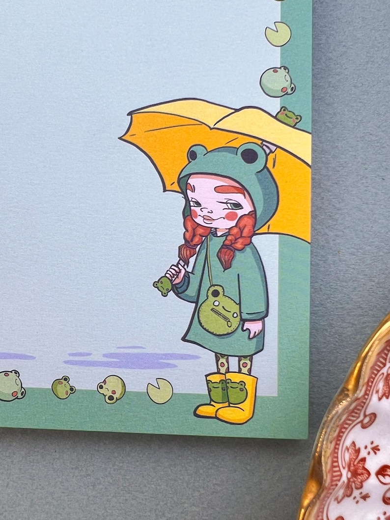 May include: A girl wearing a green frog raincoat and yellow rain boots holds a yellow umbrella. She has red hair in braids and is carrying a green frog purse. Green frogs are falling from the sky.
