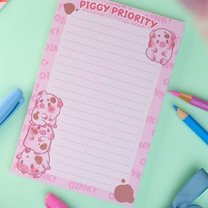 Puede incluir: Bloc de notas rosa con el texto "PIGGY PRIORITY" e ilustraciones de cerdos. El bloc de notas tiene p&aacute;ginas rayadas y est&aacute; rodeado de bol&iacute;grafos, un lazo y un bot&oacute;n sobre una superficie verde claro.