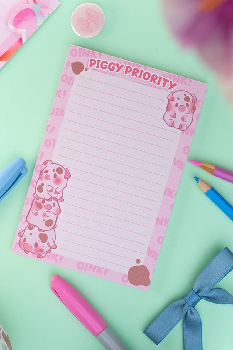 Puede incluir: Bloc de notas rosa con el texto "PIGGY PRIORITY" e ilustraciones de cerdos. El bloc de notas tiene p&aacute;ginas rayadas y est&aacute; rodeado de bol&iacute;grafos, un lazo y un bot&oacute;n sobre una superficie verde claro.