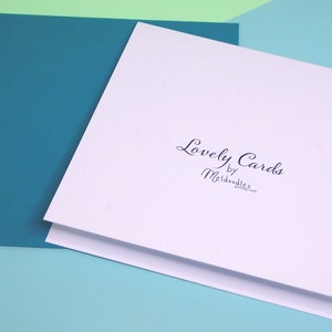 Puede incluir: Una tarjeta blanca con el texto "Lovely Cards by Meldoodles" impreso en tinta negra. La tarjeta est&aacute; sobre una superficie azul claro.