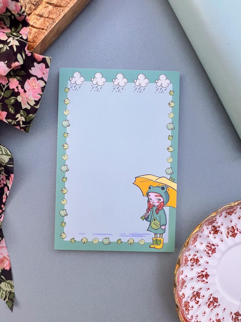 May include: A light blue notepad with a green border featuring a cartoon frog wearing a raincoat and holding an umbrella. The border is decorated with small green frogs and clouds with raindrops.