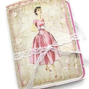 May include: A vintage-style journal with a pink cover featuring a retro illustration of a woman in a pink dress. The journal is tied with a white lace ribbon. The pages have a vintage map design.