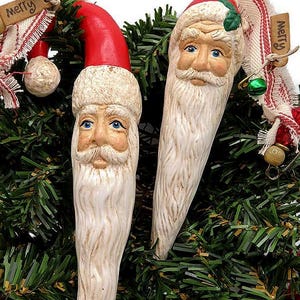 May include: Two Santa Claus Christmas ornaments with red hats and long white beards. The ornaments are set against a backdrop of green garland and gold ornaments. Each ornament has a tag that says "Merry".