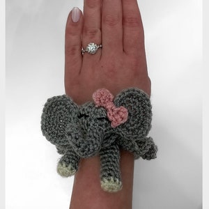 May include: A gray crocheted elephant bracelet with a pink bow. The elephant has small, closed eyes and light-colored feet. The bracelet is worn on a wrist.