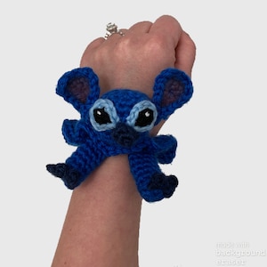 May include: Blue crocheted Stitch character from the Disney movie Lilo & Stitch. The character is wearing a blue and white wristband and has large blue eyes.