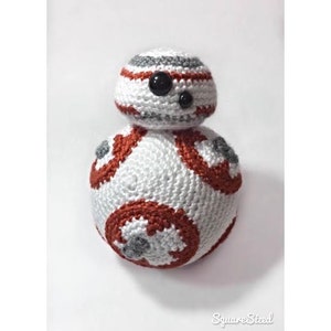May include: A white and orange crocheted toy robot with black eyes. The robot has a round body with a smaller round head on top. The robot is a popular character from the Star Wars franchise.