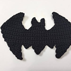 May include: A black crochet bat with outstretched wings. The handmade bat has a textured surface and a detailed design, perfect for Halloween decorations or craft projects. The bat is displayed on a white background.