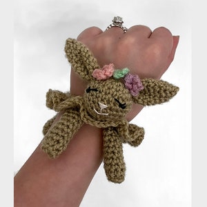 Könnte beinhalten: Ein gehäkeltes braunes Häschen mit einer rosa, grünen und hellrosa Blütenkrone. Das Häschen hat eine weiße Nase und schwarze Augen. Das Häschen ist an einem Handgelenk befestigt.