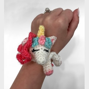 May include: A white crocheted unicorn with a pink mane and a yellow horn. The unicorn has a blue and pink flower crown and closed eyes.