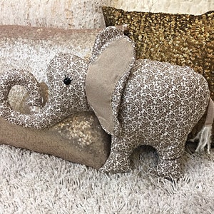 May include: A plush elephant toy with a brown and white floral pattern. The elephant is lying on a white shag rug with two pillows behind it. One pillow is a light brown color and the other is gold with sequins.
