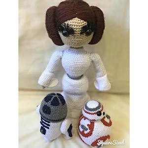 May include: Crocheted Star Wars characters, including Princess Leia, BB-8, and a droid resembling R2-D2. The characters are made with white, brown, gray, orange, and blue yarn.