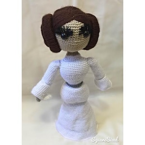 May include: A crocheted doll of Princess Leia from Star Wars. The doll is wearing a white dress and has brown hair with a white headband. The doll has black eyes with eyelashes.