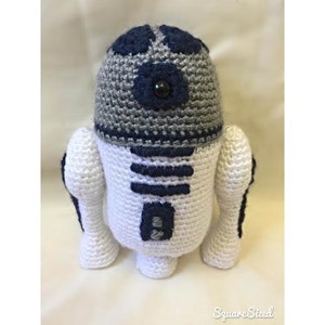 May include: A crocheted R2-D2 robot toy with a white body, blue and gray details, and a black eye.