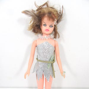 May include: A vintage doll with brown hair adorned with feathers and a silver glitter dress. The doll has blue eyes, pink lips, and a beaded necklace. A green belt and a small green accessory complete the look. The doll is standing.