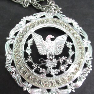 May include: Silver-toned pendant necklace featuring a detailed eagle design within a circular frame. The frame is adorned with clear rhinestones and decorative flourishes. The necklace has a matching silver chain.