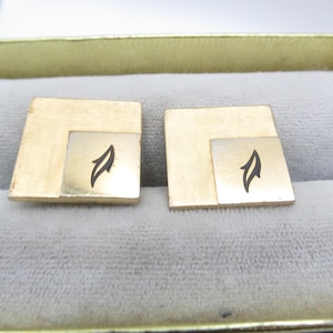 May include: A pair of gold-coloured square cufflinks. Each cufflink has a textured, larger square with a smaller, lighter square inset. A black, stylised flame design is centred on the inset square. The cufflinks are displayed in a velvet-lined box.