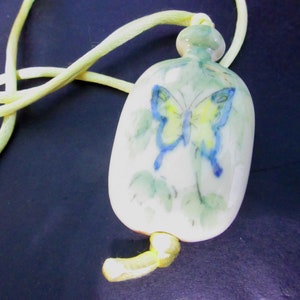 May include: A ceramic pendant necklace featuring a light green and white butterfly design. The pendant is attached to a yellow cord.