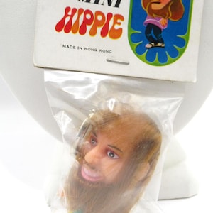 May include: A vintage "Mini Hippie" doll head in a plastic bag, with a smiling face and long brown hair and beard. The packaging features a cartoon hippie illustration and the text "Mini Hippie" and "Made in Hong Kong".