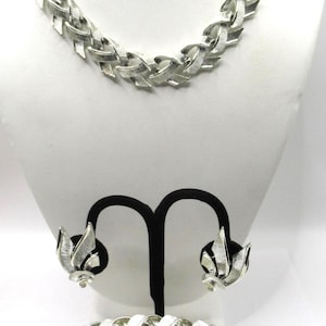 Puede incluir: Un conjunto de joyería en tono plateado que consta de un collar, una pulsera y un par de pendientes de clip. El collar y la pulsera presentan un patrón de eslabones tejidos y texturizados. Los pendientes tienen un diseño de hojas.