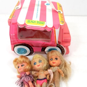 May include: A vintage pink and white striped Beach Buggy toy with three small dolls. The Beach Buggy has a handle and clear plastic windows. The dolls have blonde hair and are wearing various outfits.