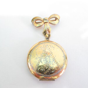 May include: A vintage gold-toned locket pendant with a bow-shaped brooch. The round locket features an etched design and opens to reveal space for a photo. The bow and locket show signs of age.