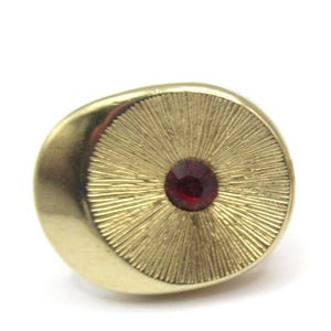 May include: A gold-coloured ring with a textured, sunburst design. It has a round, deep red gemstone in the centre. The ring is oval-shaped and set against a white background.