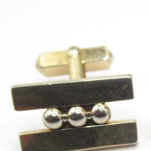May include: Gold-coloured cufflinks with a minimalist design. The cufflinks feature two rectangular bars connected by three small, silver-coloured spheres. The post and clasp are also gold-coloured, creating a cohesive look.