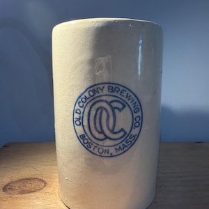 May include: A cylindrical, off-white stoneware crock with a blue circular emblem that reads "OLD COLONY BREWING CO. BOSTON, MASS." The crock has a slightly textured surface and a crack near the top. It is a vintage piece, likely used for brewing or storage.
