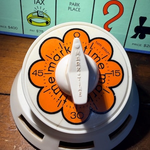 May include: A white and orange Mark Time kitchen timer with a dial showing numbers 15, 30, and 45. The timer has a white knob and the words "mark time" are printed on the dial. A Monopoly board is in the background.