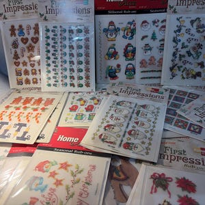 May include: A collection of First Impressions rub-on transfer sheets featuring various Christmas and winter-themed designs. The sheets include images of gingerbread men, snowmen, candy canes, and other festive elements. The packaging is white and red.