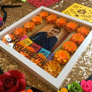 May include: A white picture frame with a photo of a man inside. The frame is decorated with orange paper flowers and colorful paper cutouts. The text "Recuerdame" is written in white on the frame.
