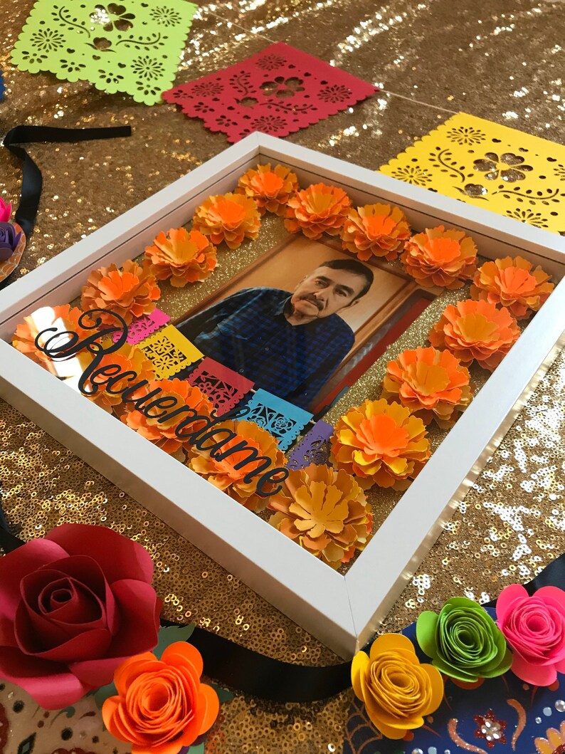 May include: A white picture frame with a photo of a man inside. The frame is decorated with orange paper flowers and colorful paper cutouts. The text "Recuerdame" is written in white on the frame.