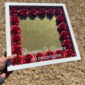 May include: A white frame with a gold glitter background surrounded by red paper roses. The frame has the text "Siempre te llevaré en mi corazón" written in white.