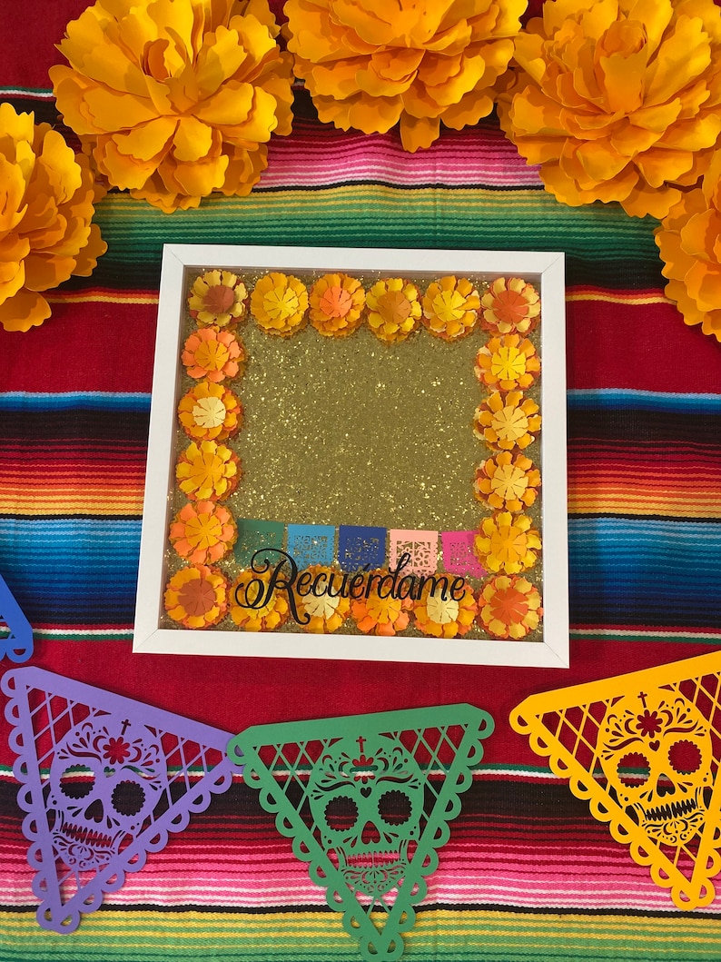 May include: A white frame with a gold glitter background and a border of orange paper flowers. The frame has the text "Recu&eacute;rdame" written in black cursive. The frame is surrounded by orange paper flowers and a colorful Mexican blanket.