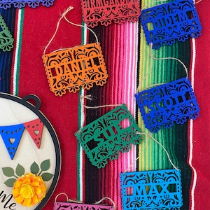 Puede incluir: Un conjunto de diez banderines de papel de colores con nombres escritos en ellos. Los banderines están decorados con intrincados diseños recortados y están unidos entre sí con un cordel. Los banderines están colgados de un fondo de tela de rayas de colores.