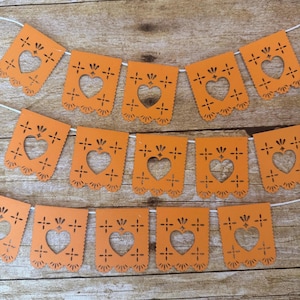 Puede incluir: Una guirnalda decorativa de papel naranja con un patrón repetitivo de corazones y motivos florales troquelados. La guirnalda consta de varios banderines rectangulares unidos por un cordel blanco, con bordes festoneados en la parte inferior.