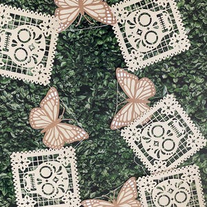 May include: A decorative banner featuring cream-colored, cut-out skull and floral designs, interspersed with tan butterfly cutouts. The banner is strung together with white string, perfect for festive occasions.