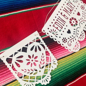 May include: White paper banner with cut-out designs, strung with white string. The banner reads "EDWIN & MARY" in red lettering. The banner is hanging on a colorful striped fabric.