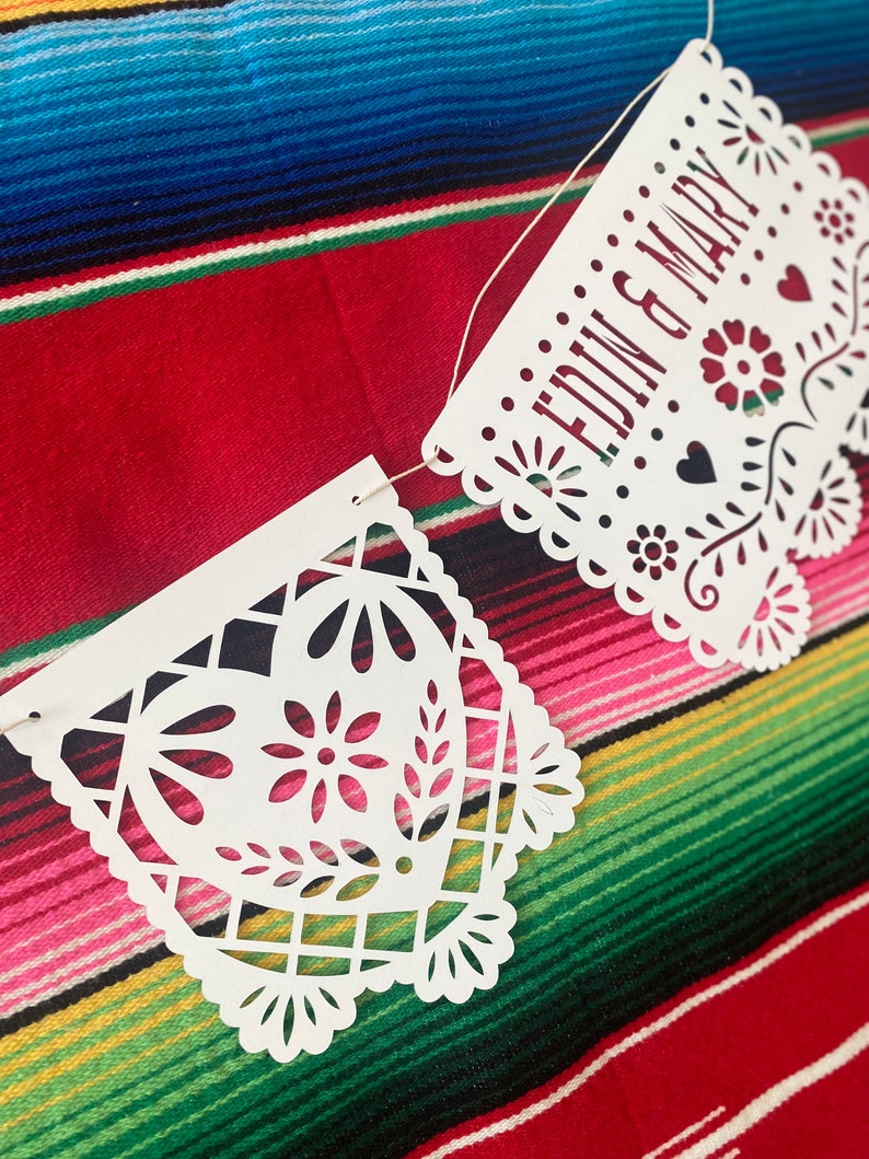 May include: White paper banner with cut-out designs, strung with white string. The banner reads "EDWIN & MARY" in red lettering. The banner is hanging on a colorful striped fabric.