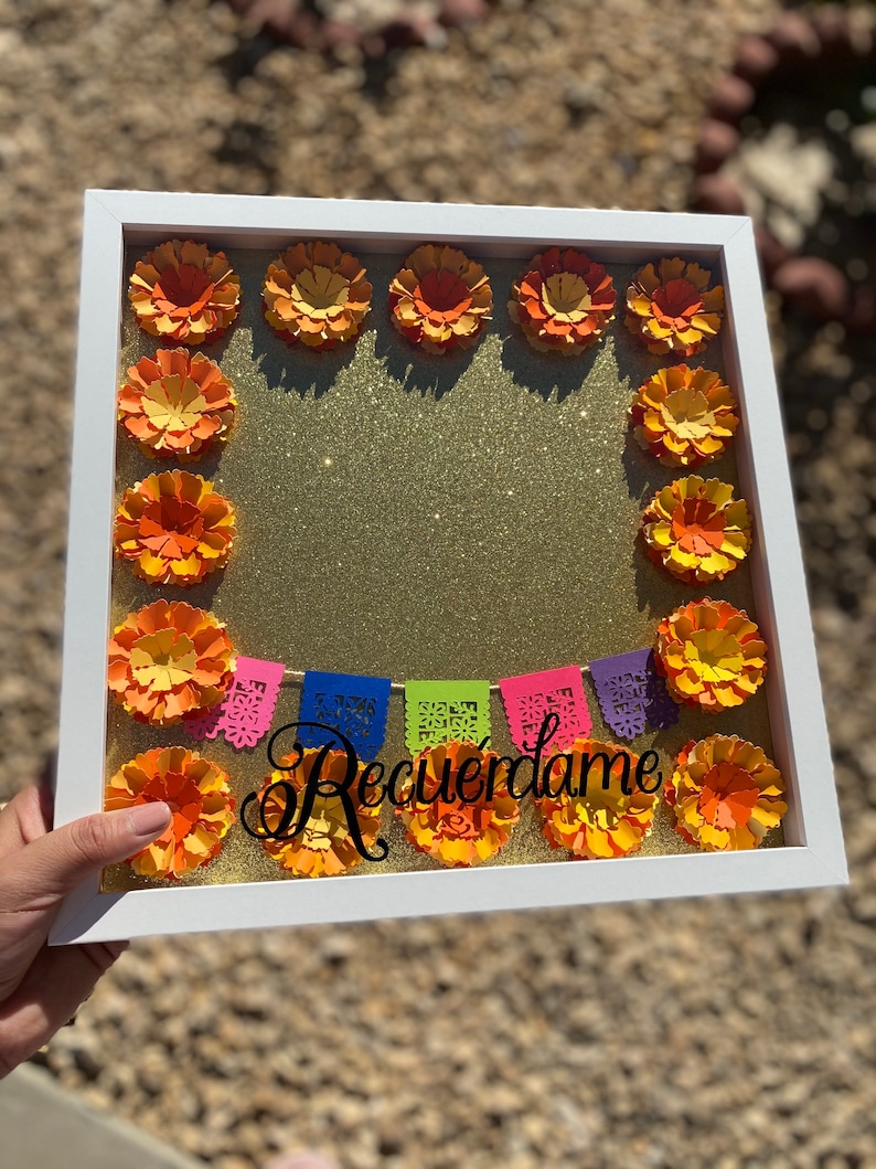 May include: A white picture frame with a gold glitter background. The frame is decorated with orange paper flowers and a colorful paper banner that reads "Recu&eacute;rdame".