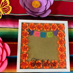 May include: A white frame with a gold glitter background surrounded by orange paper flowers. The frame has the word "Fortaleza" written in black cursive lettering. There are colorful paper cutouts hanging from the top of the frame.