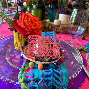 May include: A pink paper place card with the name "KELLI" cut out in a decorative font. The place card is sitting on a small white bowl with colorful pom-poms around the rim. The bowl is on a blue and white patterned fabric with tassels. The fabric is on a clear glass plate with a purple lace doily underneath.